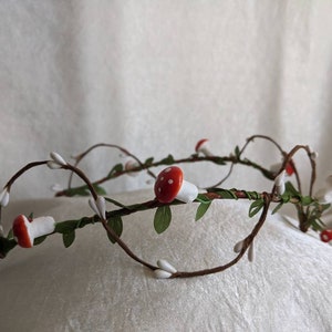 May include: A crown made of twigs and branches with small white flowers and red and white mushroom decorations.