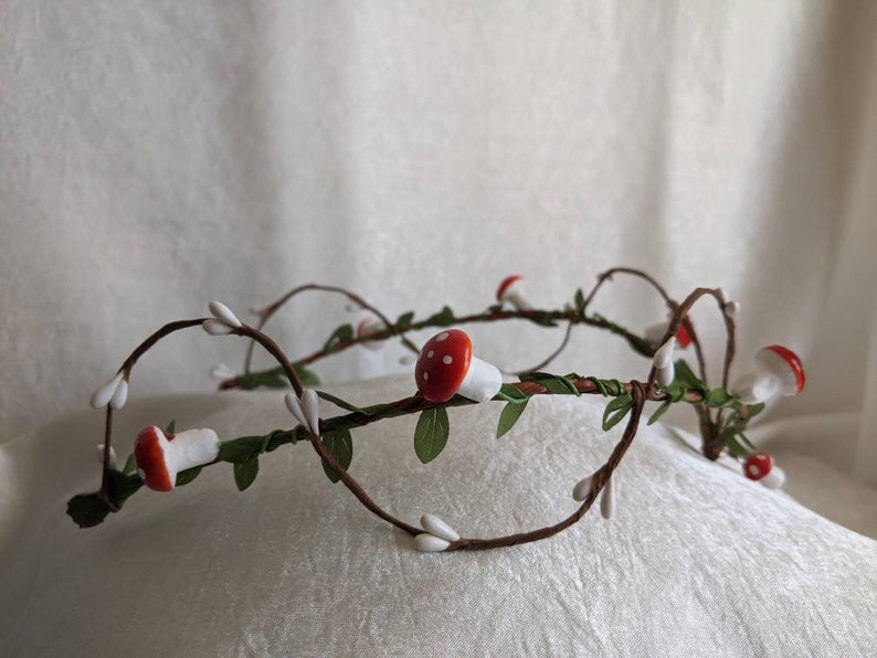 May include: A crown made of twigs and branches with small white flowers and red and white mushroom decorations.