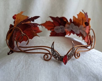 Può includere: Una corona in filo di rame con un design a mezzaluna, ornata da una gemma rossa e foglie autunnali. Le foglie sono nelle tonalità dell'arancione, del rosso e del marrone, con piccole bacche rosse. La corona poggia su una superficie bianca.