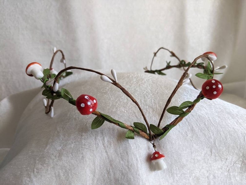 May include: A woodland-themed crown made of brown twigs, green leaves, and white and red mushroom-shaped ornaments. The crown has a dangling red and white mushroom ornament.
