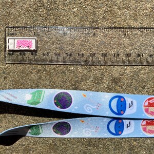 May include: A light blue lanyard with a repeating pattern of cartoon food and objects. The pattern includes a green box with a white lid, a purple flower, a white cloud with a star, a blue bucket with a white lid and a cartoon face, and a pink and red bowl with chopsticks. The lanyard is approximately 1 inch wide.