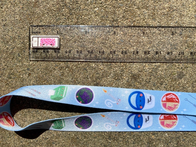 May include: A light blue lanyard with a repeating pattern of cartoon food and objects. The pattern includes a green box with a white lid, a purple flower, a white cloud with a star, a blue bucket with a white lid and a cartoon face, and a pink and red bowl with chopsticks. The lanyard is approximately 1 inch wide.
