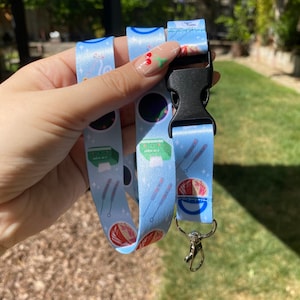 May include: A blue lanyard with a white and pink pattern featuring various illustrations of yarn, needles, and a bowl of yarn. The lanyard has a black plastic clip and a silver metal clasp.
