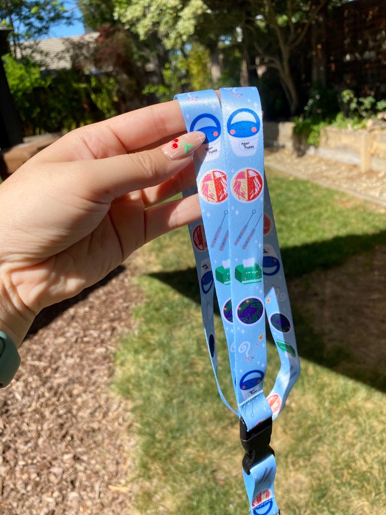May include: A blue lanyard with a white background and colorful illustrations of science-related items, including petri dishes, test tubes, and a beaker. The text "Agar Plates" is visible on the lanyard.