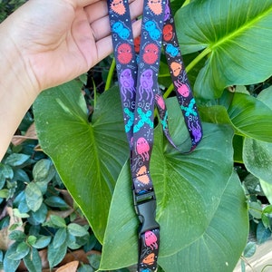 Peut inclure: Lanière noire ornée d'un motif répété de microbes de dessins animés colorés et du texte "Bio 8". Elle est dotée d'un fermoir en plastique noir et d'un clip en métal argenté. Le fond est un feuillage vert.