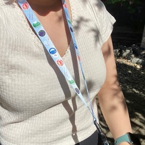 May include: A white shirt with a blue and white lanyard with various colorful icons. The lanyard has a black clip at the bottom.