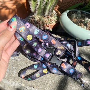 Peut inclure: Un cordon noir avec un motif coloré de microbes de dessin animé, de brins d'ADN et de symboles scientifiques. Le cordon a un clip en plastique noir et un crochet en métal argenté.