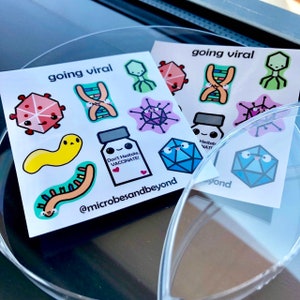 Op de afbeelding: Twee vellen stickers met cartoon illustraties van virussen, DNA en vaccins. Tekst bevat "going viral" en "Don't Hesitate, VACCINATE!". De stickers liggen op een doorzichtig oppervlak.