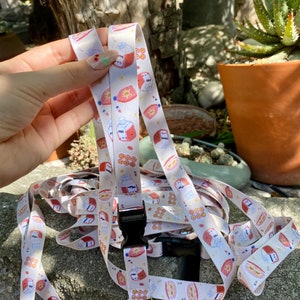 May include: A white lanyard with a repeating pattern of cartoon food illustrations in red, orange, and blue. The lanyard has a black plastic clasp and a gold-colored metal swivel hook. The lanyard is made of a smooth, satin-like material.