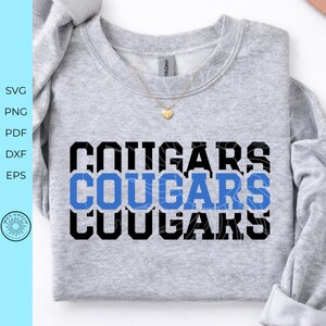 May include: A light grey crewneck sweatshirt featuring the word COUGARS printed in three stacked rows. The top and bottom rows are in black block lettering, while the middle row is in blue block lettering. A gold heart pendant necklace rests on the neckline.