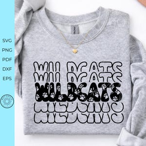 May include: A light gray crewneck sweatshirt featuring a black graphic design with the text "WILDCATS" in a bold, wavy font. The letters are filled with small paw print patterns. A gold-toned heart pendant necklace rests on the neckline.