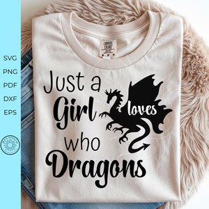 May include: A beige crew neck t-shirt featuring black text that reads Just a Girl who loves Dragons, with the word loves written in a script font. A black silhouette of a dragon is positioned between the words loves and Dragons.