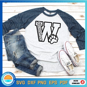 May include: A white and blue baseball tee shirt with a black graphic of a large letter W with the word "Wolverines" written inside the letter. A paw print is inside the letter W. The shirt is on a wooden surface with a pair of white sneakers and a pair of blue jeans.