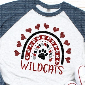 Op de afbeelding: Een baseball-stijl T-shirt in gem&ecirc;leerd grijs met donkerblauwe mouwen, voorzien van een regenboogafbeelding met rode, zwarte en grijze strepen, witte pootafdrukken en rode hartjes. Het woord WILDCATS staat in het rood onder de regenboog gedrukt.
