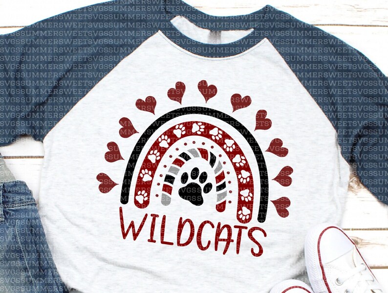Op de afbeelding: Een baseball-stijl T-shirt in gem&ecirc;leerd grijs met donkerblauwe mouwen, voorzien van een regenboogafbeelding met rode, zwarte en grijze strepen, witte pootafdrukken en rode hartjes. Het woord WILDCATS staat in het rood onder de regenboog gedrukt.