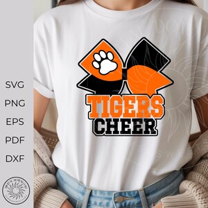 Pode incluir: Camiseta branca com um la&ccedil;o laranja e preto com uma pegada branca. A frase "TIGERS CHEER" est&aacute; impressa em laranja e preto. A camiseta &eacute; usada por uma pessoa.