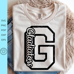 May include: A light beige short-sleeved t-shirt featuring a large black varsity-style letter G with the word Gladiators written in a black script font vertically along the left side of the letter. The shirt has a crew neckline and a visible brand tag.