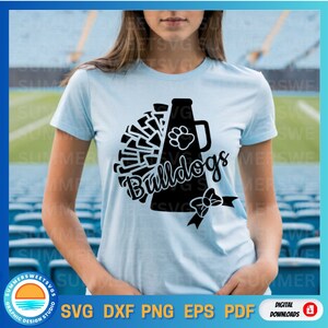 May include: Light blue t-shirt featuring a black graphic of a megaphone, pom-poms, and the word "Bulldogs." A paw print and bow are also included in the design. The shirt is a digital download.