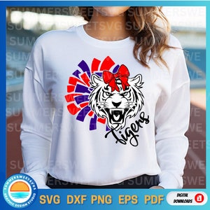Puede incluir: Sudadera blanca con un pompón rojo, blanco y azul y una cabeza de tigre blanco y negro con un lazo rojo. El texto "Tigers" está escrito en cursiva negra debajo de la cabeza del tigre.