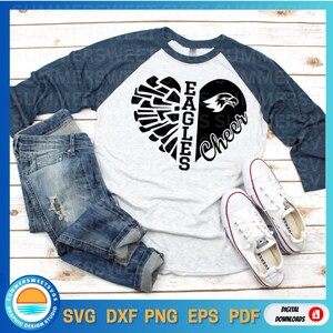 Puede incluir: Una camiseta de b&eacute;isbol blanca y azul con un dise&ntilde;o de coraz&oacute;n negro. El coraz&oacute;n est&aacute; dividido en dos, con el lado izquierdo que presenta un ala de &aacute;guila estilizada y el lado derecho que presenta la palabra "Cheer". El texto "Eagles" est&aacute; escrito verticalmente en el lado izquierdo del coraz&oacute;n.