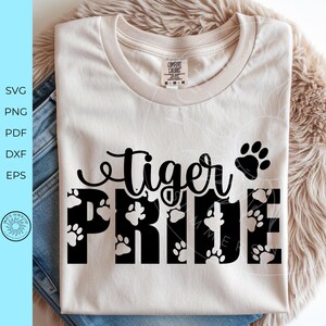 May include: A cream-colored t-shirt featuring the black text tiger PRIDE with a paw print icon. The word tiger is in a cursive script, while PRIDE is in a bold, block font filled with small white paw prints. The shirt has a Comfort Colors label.