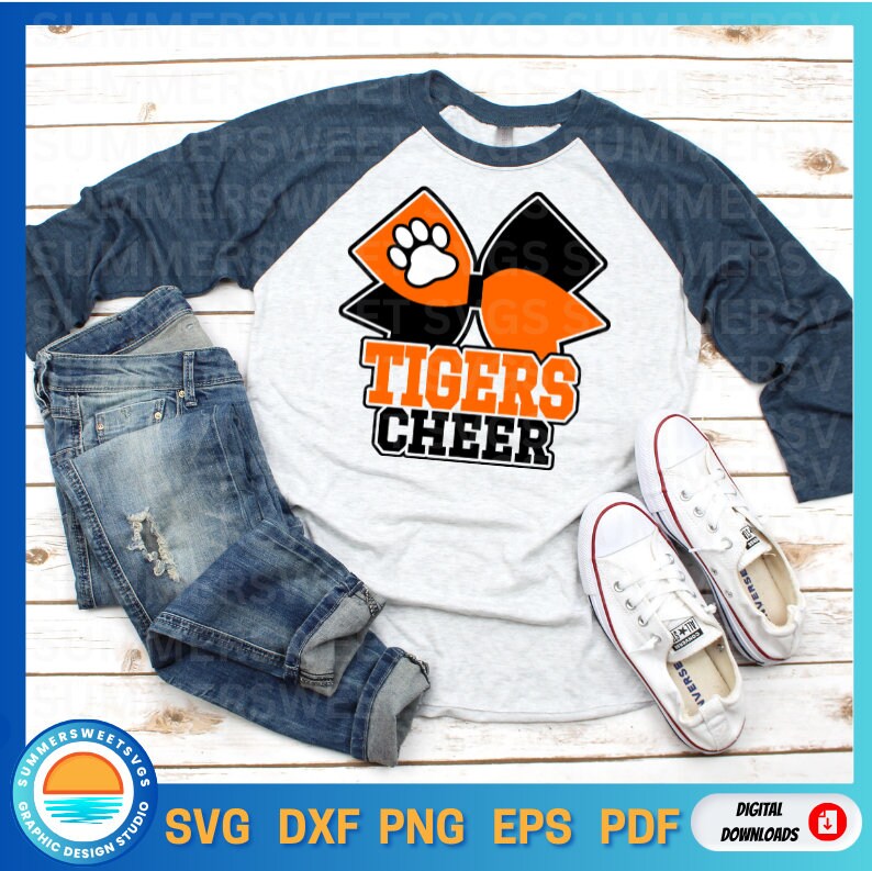 Pode incluir: Uma camisa cinza e azul marinho estilo raglan com as palavras "TIGERS CHEER" em laranja e preto. A camisa apresenta um gr&aacute;fico de la&ccedil;o com pegada de pata. A imagem tamb&eacute;m inclui um par de jeans azuis e t&ecirc;nis brancos.