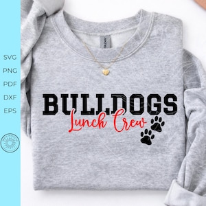 May include: A heather gray crewneck sweatshirt featuring the black text BULLDOGS in a bold block font, with the red script text Lunch Crew underneath, accompanied by two black paw prints. A gold heart-shaped pendant necklace rests at the neckline.