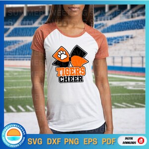 Pode incluir: Camiseta raglan branca e laranja com um gr&aacute;fico de la&ccedil;o preto e laranja. O la&ccedil;o apresenta uma pegada e as palavras "TIGERS CHEER" em laranja e preto. A camiseta &eacute; um download digital.