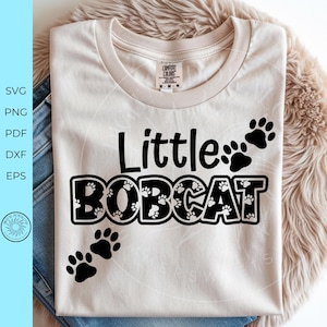 May include: Cream-colored t-shirt with the words "Little BOBCAT" in black, with paw prints. The shirt is laid on top of blue jeans and a fluffy, light brown surface. The shirt has a tag that says "COMFORT COLORS".