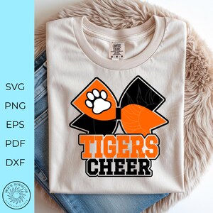 Pode incluir: Camiseta creme com um grande la&ccedil;o laranja e preto, uma pegada branca e as palavras "TIGERS CHEER" em laranja e preto. A camisa &eacute; exibida em uma superf&iacute;cie fofa marrom claro com um par de jeans.