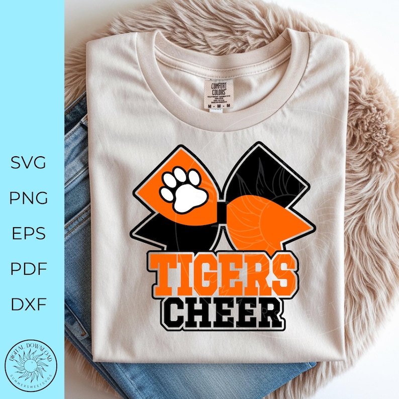 Pode incluir: Camiseta creme com um grande la&ccedil;o laranja e preto, uma pegada branca e as palavras "TIGERS CHEER" em laranja e preto. A camisa &eacute; exibida em uma superf&iacute;cie fofa marrom claro com um par de jeans.