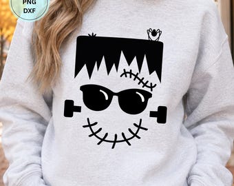 May include: A light gray crewneck sweatshirt featuring a black graphic of a monster face with jagged hair, sunglasses, a stitched forehead scar, a stitched mouth, and bolts on the sides. A small circle in the corner contains the text SVG PNG DXF.