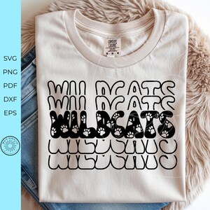 May include: A cream-colored t-shirt featuring the word WILDCATS repeated five times in a stacked, wavy font. The middle instance of the word is filled with a black pattern of small white paw prints, while the other four lines are outlined in black.