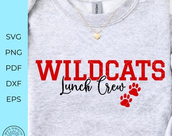 Puede incluir: Una sudadera gris claro de cuello redondo con el texto rojo WILDCATS en fuente de bloque, seguido del texto negro en cursiva Lunch Crew y dos huellas de pata rojas. Un collar con colgante en forma de corazón dorado descansa en el cuello.