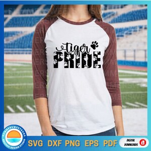 May include: A white raglan t-shirt with maroon three-quarter length sleeves. The front features the black text "tiger PRIDE" with a paw print icon. The word "PRIDE" is filled with a repeating pattern of small white paw prints.