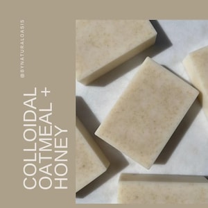 May include: Several rectangular bars of soap with a light beige color and speckled texture are arranged on a white surface. The bars are labeled "Colloidal Oatmeal + Honey" in white text on a vertical tan background.