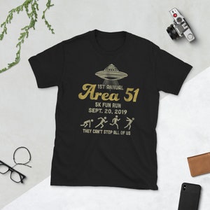 May include: Black t-shirt with a gold graphic of a UFO and the text "1st Annual Area 51 5K Fun Run Sept. 20, 2019 They Can't Stop All Of Us".