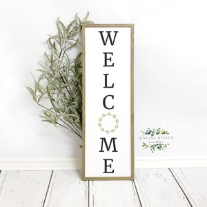 Puede incluir: Un letrero de madera con la palabra "WELCOME" escrita verticalmente en letras negras. Una pequeña corona verde se encuentra entre la "C" y la "M". El letrero está sobre un suelo de madera blanca con plantas verdes en el fondo.