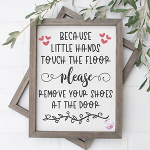 May include: A white sign with black text that reads "Because little hands touch the floor please remove your shoes at the door". The sign is framed in a light brown wood frame and has a small floral design in the bottom right corner.