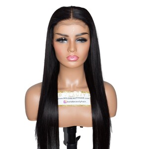 May include: A mannequin head displaying a long, straight, black wig. The wig has a natural-looking hairline and is styled with a center part. The mannequin has makeup applied. A gold sticker with the website address and Instagram handle is visible.