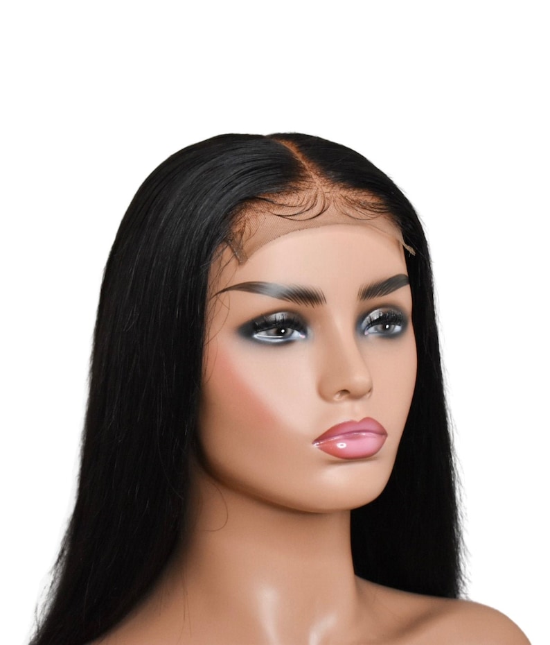 May include: A black, long, straight hair wig on a mannequin head. The wig features a natural-looking hairline and is styled with a center part. The mannequin has a fair complexion, with makeup including dark eyeshadow and pink lipstick.