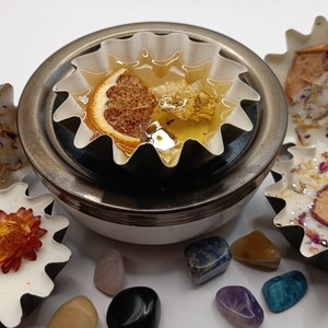 May include: A white ceramic wax warmer with a metal base, holding a white wax melt with dried orange slices and flowers.  Surrounding the warmer are various colorful gemstones.
