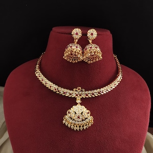 May include: A gold-toned necklace and earring set with red and clear gemstones. The necklace has a pendant with a floral design, and the earrings are bell-shaped. The jewelry is displayed on a red bust against a black background.