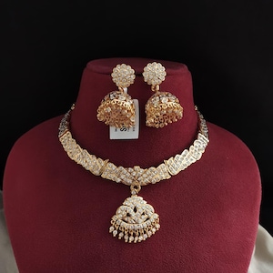 Op de afbeelding: Een goudkleurige ketting- en oorbellenset met heldere stenen. De ketting heeft een brede, gestructureerde band met een centrale hanger en bungelende kralen. De bijpassende oorbellen hebben een bloemvormige bovenkant en een klokvormige basis.