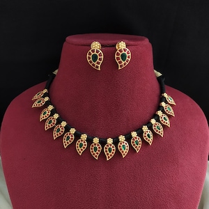 May include: A gold-coloured necklace and matching earrings set. The jewellery features a leaf-shaped design with green and red accents. The necklace is strung on a black cord, and the earrings have a similar design. The set is displayed on a red bust.