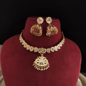 May include: A gold-toned jewelry set featuring a necklace and matching earrings. The necklace has a floral pendant with white and red accents, and the earrings are bell-shaped with similar detailing. The set is displayed on a red surface.