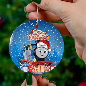 May include: Circular Christmas decoration with a cartoon train wearing a Santa hat, bearing the name "Daniel". The ornament has a blue backdrop with falling snow and the words "Merry Christmas".