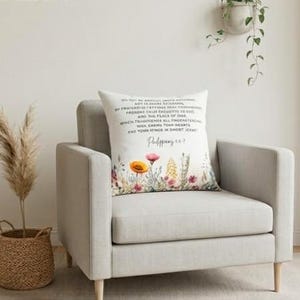 Puede incluir: Un cojín decorativo con detalles florales y texto de Filipenses 4:7. El cojín presenta un diseño de acuarela de flores coloridas en tonos rosa, amarillo y verde. El cojín está colocado sobre un sillón gris claro.