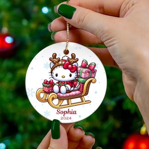 May include: A round Christmas ornament featuring a cartoon character in a red and white outfit, seated on a sleigh with gifts. The ornament has the name "Sophia" and the year "2024" printed on it. The ornament is held by a person.