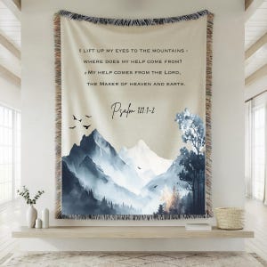 May include: A woven throw blanket with a mountain scene in blue and grey, and text from Psalm 121:1-2. The design includes birds and trees, with a fringed edge. The text reads: "I lift up my eyes to the mountains - Where does my help come from? My help comes from the Lord, the maker of heaven and earth."